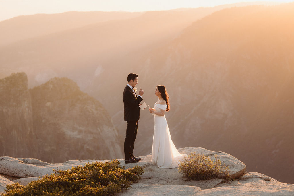 A couple stands facing each other at the edge of a cliff during sunset. The man is in a suit, and the woman is in a white dress, with mountains in the background at Taft Point 