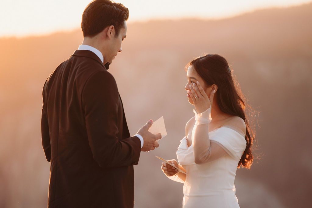 A couple stands facing each other at the edge of a cliff during sunset. The man is in a suit, and the woman is in a white dress, with mountains in the background at Taft Point 