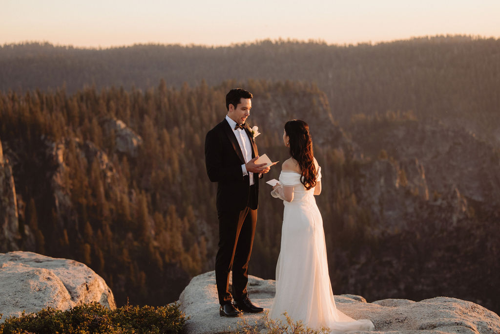 A couple stands facing each other at the edge of a cliff during sunset. The man is in a suit, and the woman is in a white dress, with mountains in the background at Taft Point 