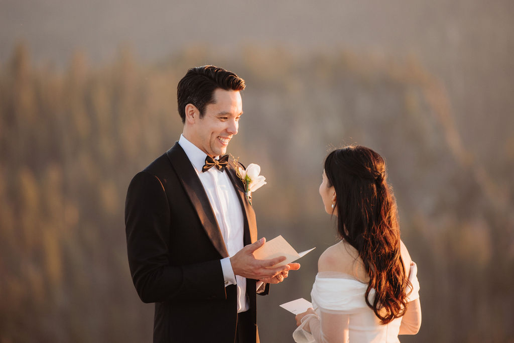 A couple stands facing each other at the edge of a cliff during sunset. The man is in a suit, and the woman is in a white dress, with mountains in the background at Taft Point 