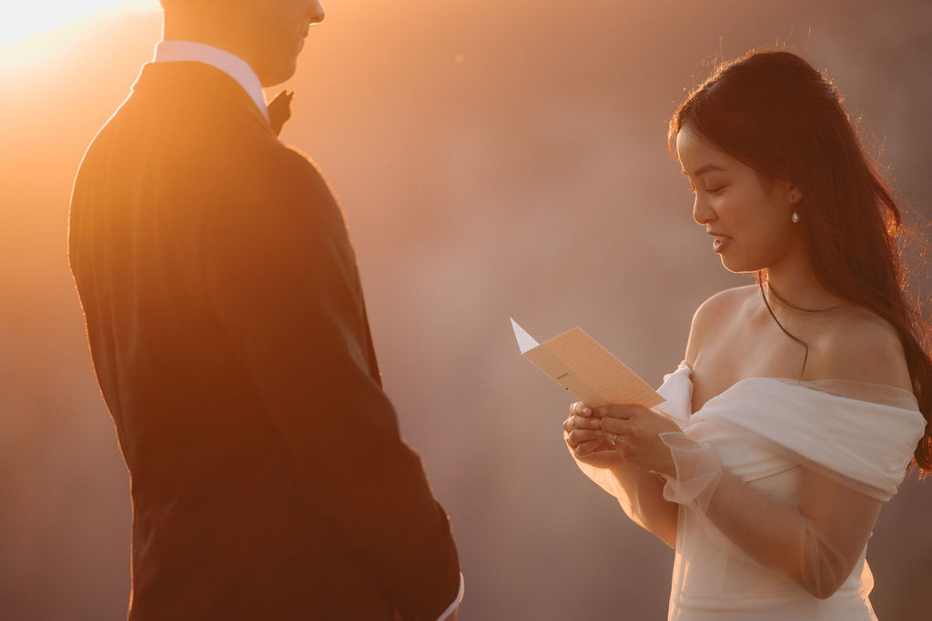 A couple stands facing each other at the edge of a cliff during sunset. The man is in a suit, and the woman is in a white dress, with mountains in the background at Taft Point 