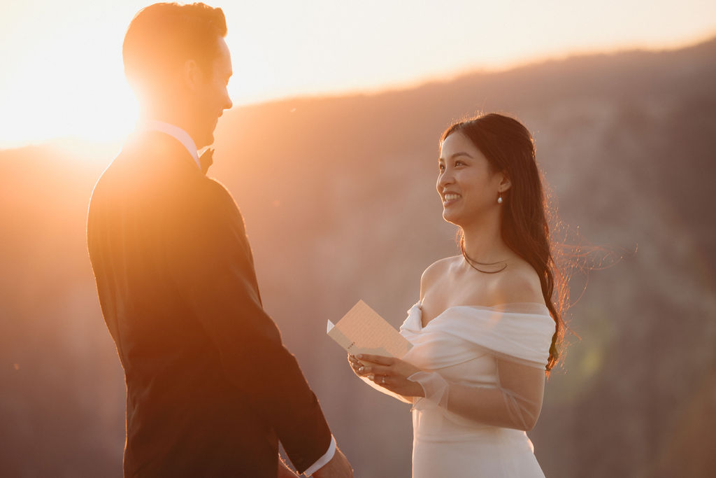 A couple stands facing each other at the edge of a cliff during sunset. The man is in a suit, and the woman is in a white dress, with mountains in the background at Taft Point 