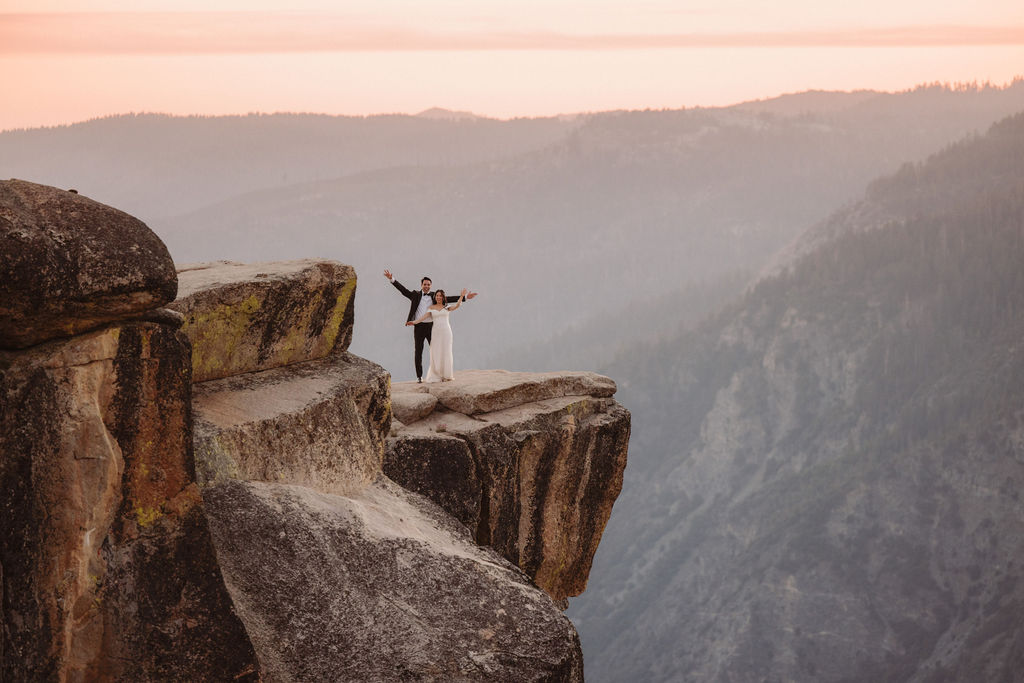 A couple stands on a rocky cliff edge with arms outstretched, overlooking a vast mountainous landscape during sunset at Taft Point