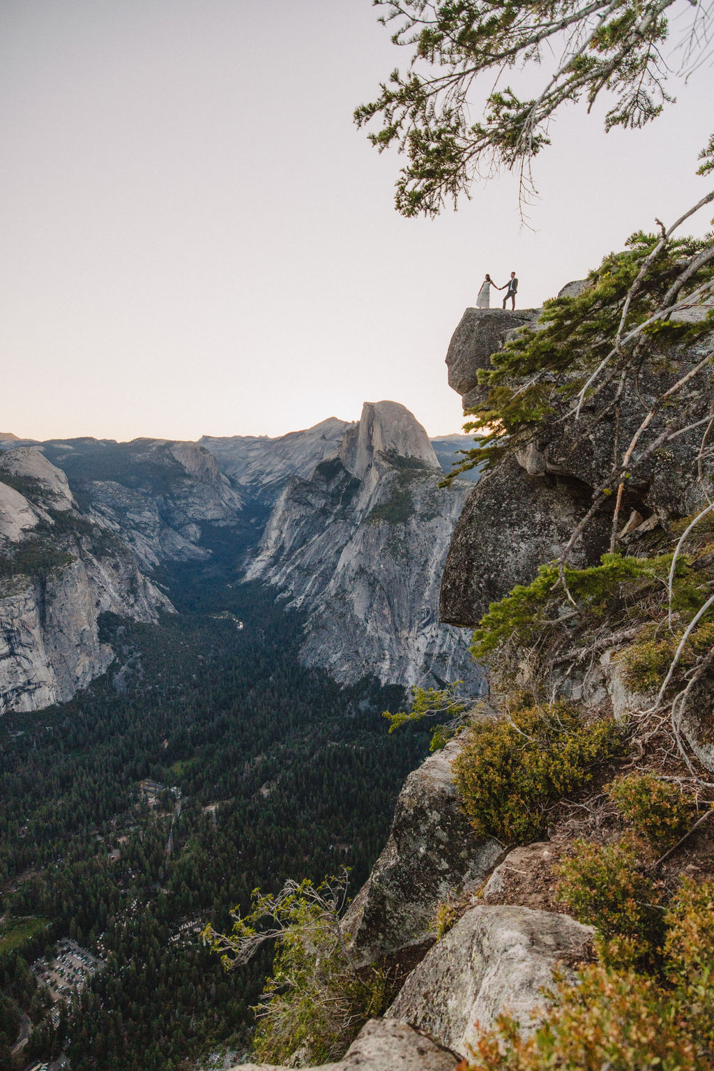 A couple, dressed in black and white, stands on a cliff edge overlooking a vast mountainous landscape, with trees and rocky cliffs in the background for their Yosemite elopement