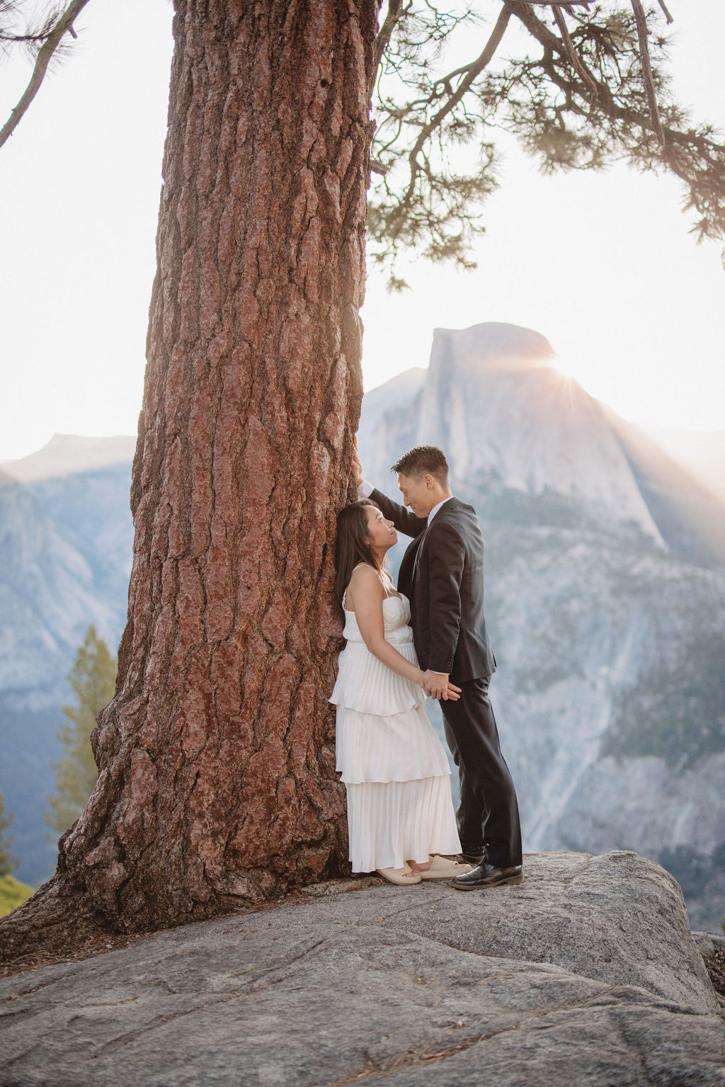 A couple stands close to each other in front of a large tree with sunlight filtering through the branches and distant mountains in the background for their Yosemite Elopement 