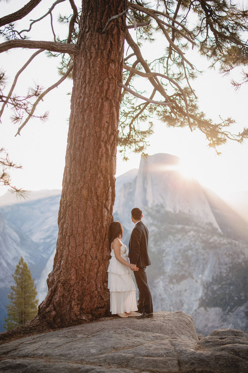 A couple stands close to each other in front of a large tree with sunlight filtering through the branches and distant mountains in the background for their Yosemite Elopement 