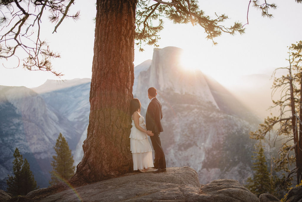 A couple stands close to each other in front of a large tree with sunlight filtering through the branches and distant mountains in the background for their Yosemite Elopement 