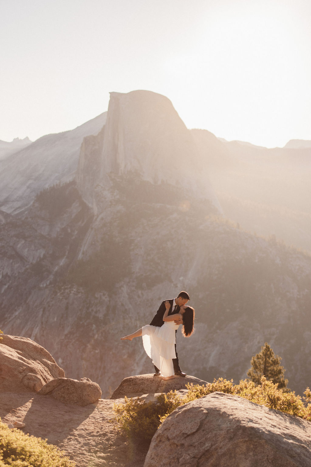 A couple holding hands stands on a rock ledge, overlooking a distant mountain peak under a hazy sky for their Yosemite elopement 