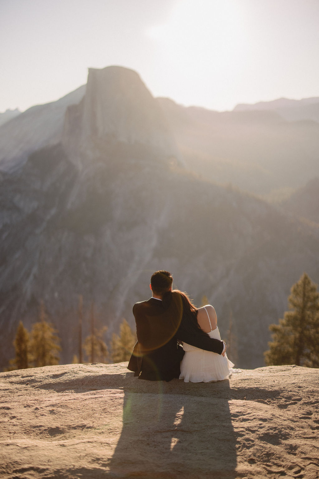 A couple holding hands stands on a rock ledge, overlooking a distant mountain peak under a hazy sky for their Yosemite elopement 