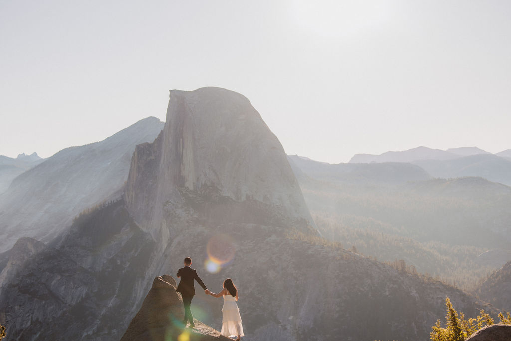 A couple holding hands stands on a rock ledge, overlooking a distant mountain peak under a hazy sky for their Yosemite elopement 