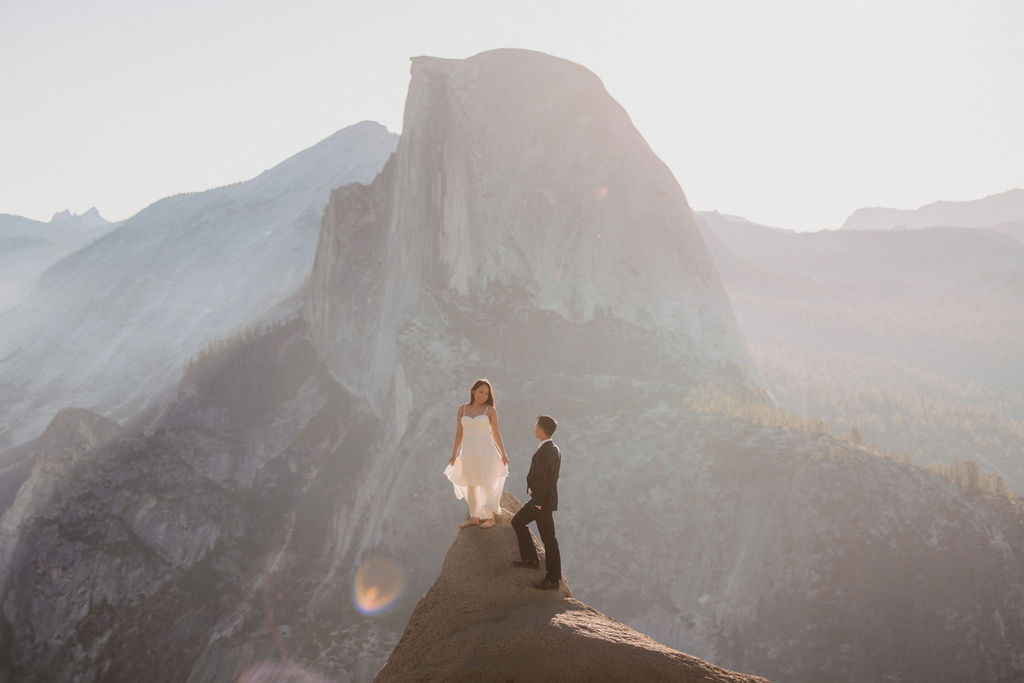 A couple holding hands stands on a rock ledge, overlooking a distant mountain peak under a hazy sky for their Yosemite elopement 