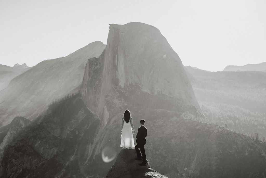 A couple holding hands stands on a rock ledge, overlooking a distant mountain peak under a hazy sky for their Yosemite elopement