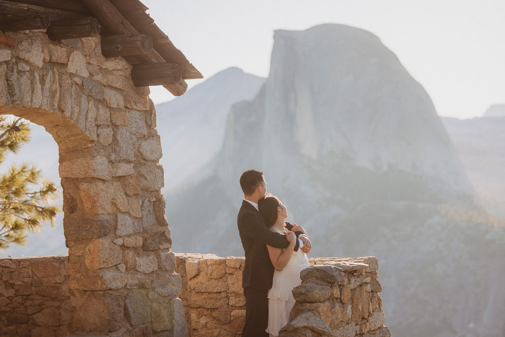 A couple in wedding attire stands on a stone balcony with a scenic view of mountains and cliffs in the background for their Yosemite elopement