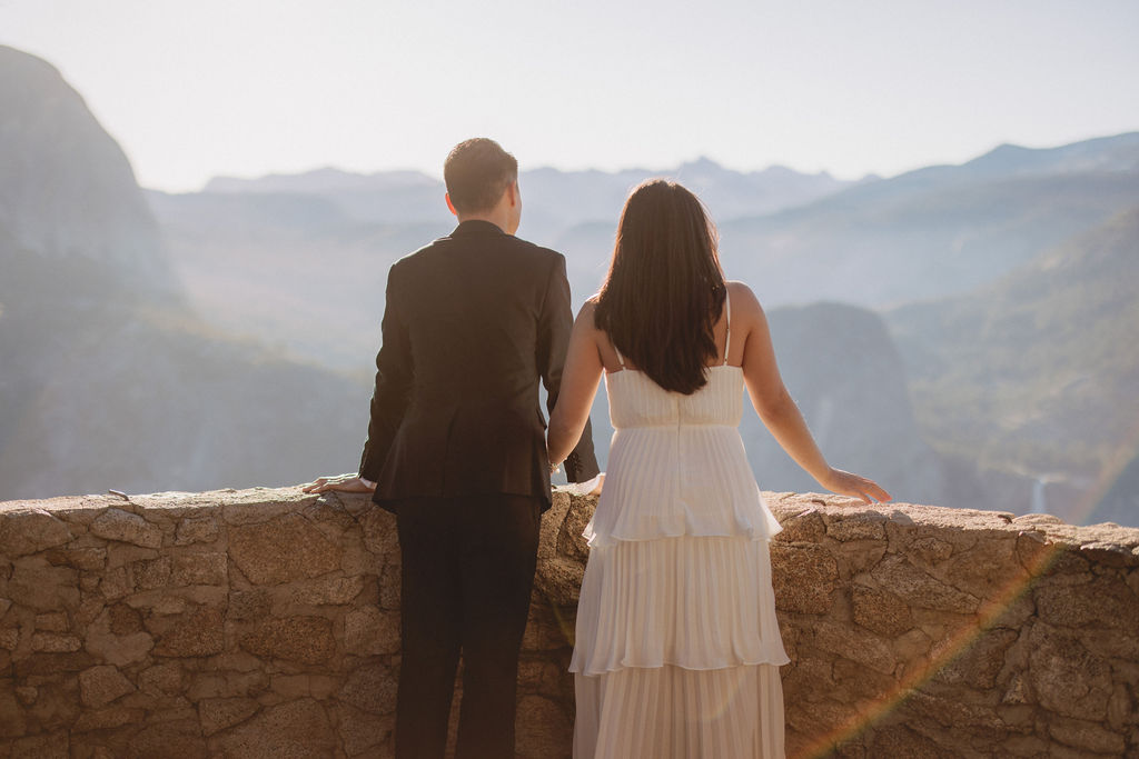 A couple in wedding attire stands on a stone balcony with a scenic view of mountains and cliffs in the background for their Yosemite elopement