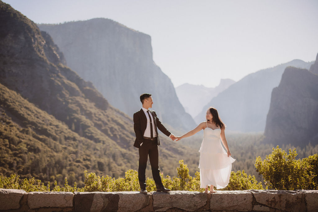 A couple in wedding attire embraces on a stone terrace with a wooden roof, overlooking a mountainous landscape illuminated by sunlight for their Yosemite elopement 