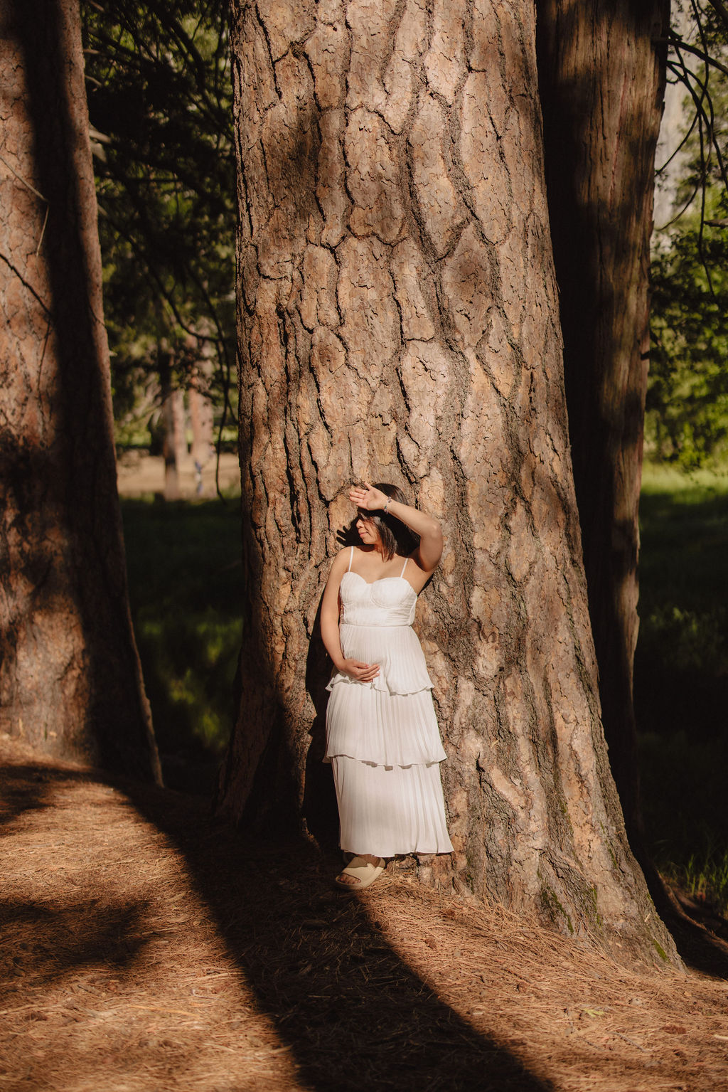 A person in a white dress stands against a large tree trunk, with one hand on a hat, in a forest setting.