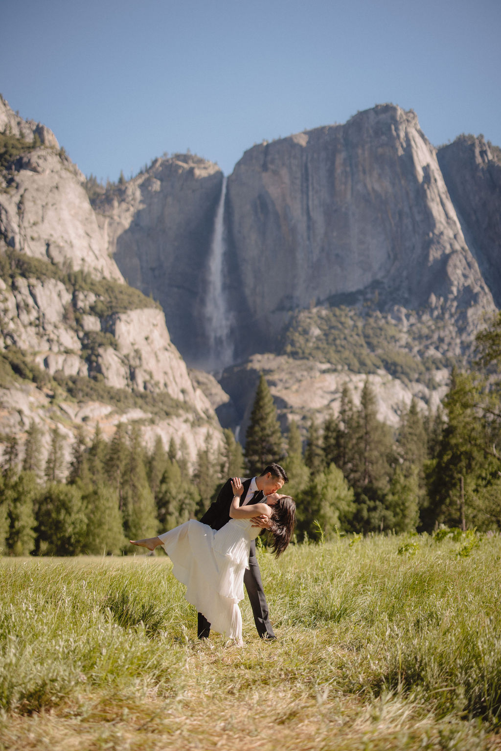 A couple in formal attire hold hands in a grassy field with a waterfall and towering cliffs in the background