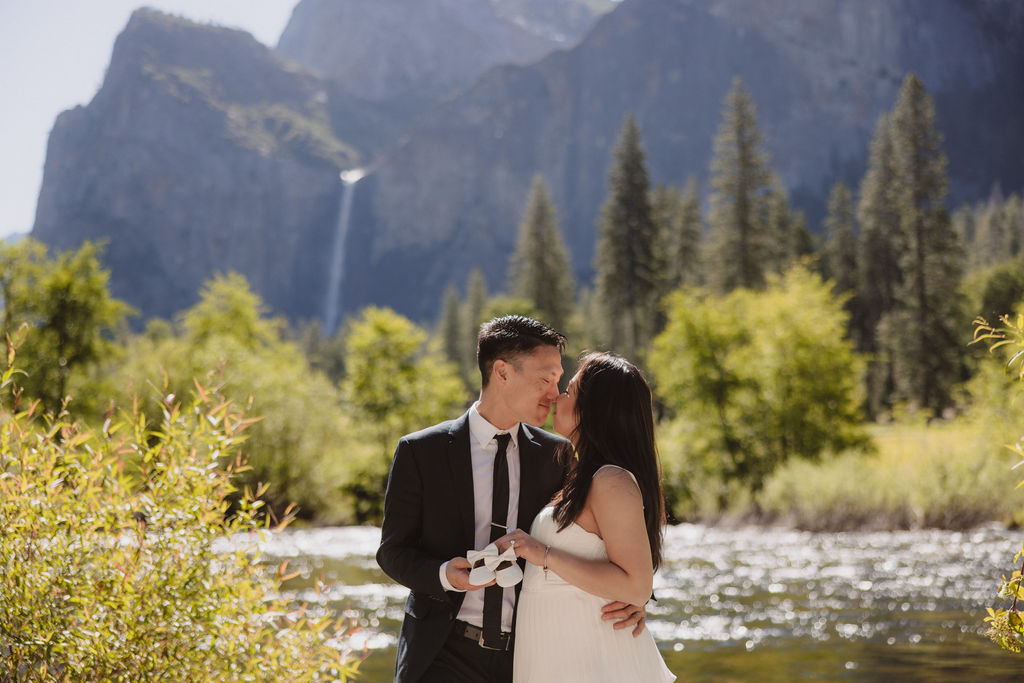 A couple in formal attire hold hands in a grassy field with a waterfall and towering cliffs in the background 