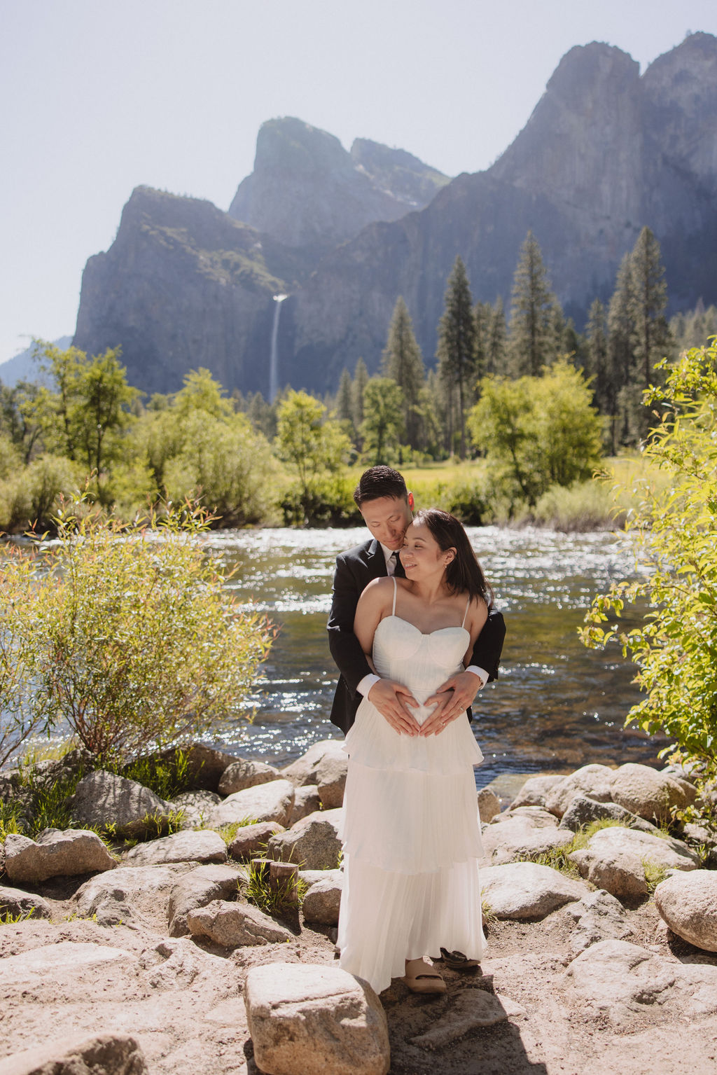 A couple in formal attire hold hands in a grassy field with a waterfall and towering cliffs in the background 