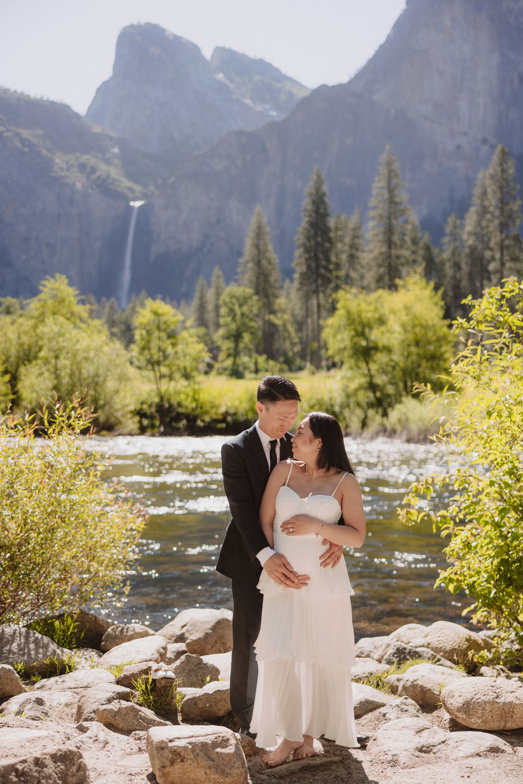 A couple in formal attire hold hands in a grassy field with a waterfall and towering cliffs in the background 