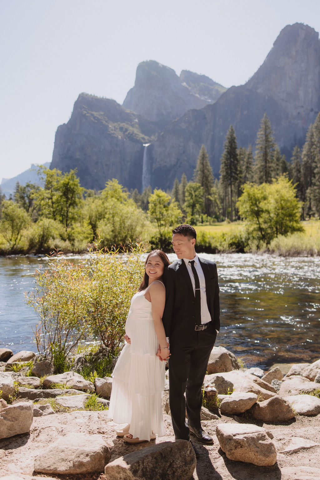 A couple in formal attire hold hands in a grassy field with a waterfall and towering cliffs in the background 