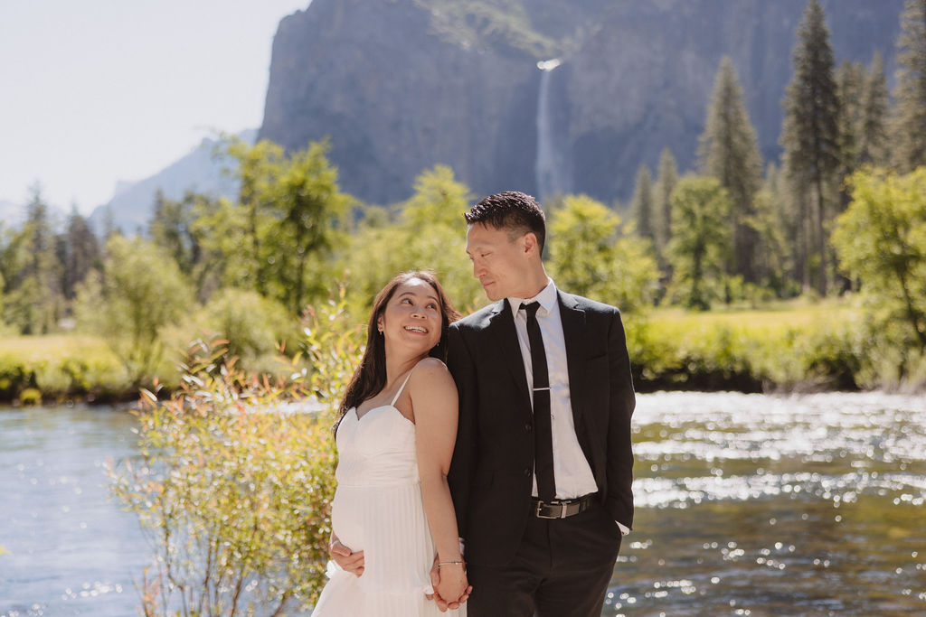 A couple, dressed formally, stands hand in hand near a river with a mountainous landscape and waterfall in the background at their Yosemite elopement 