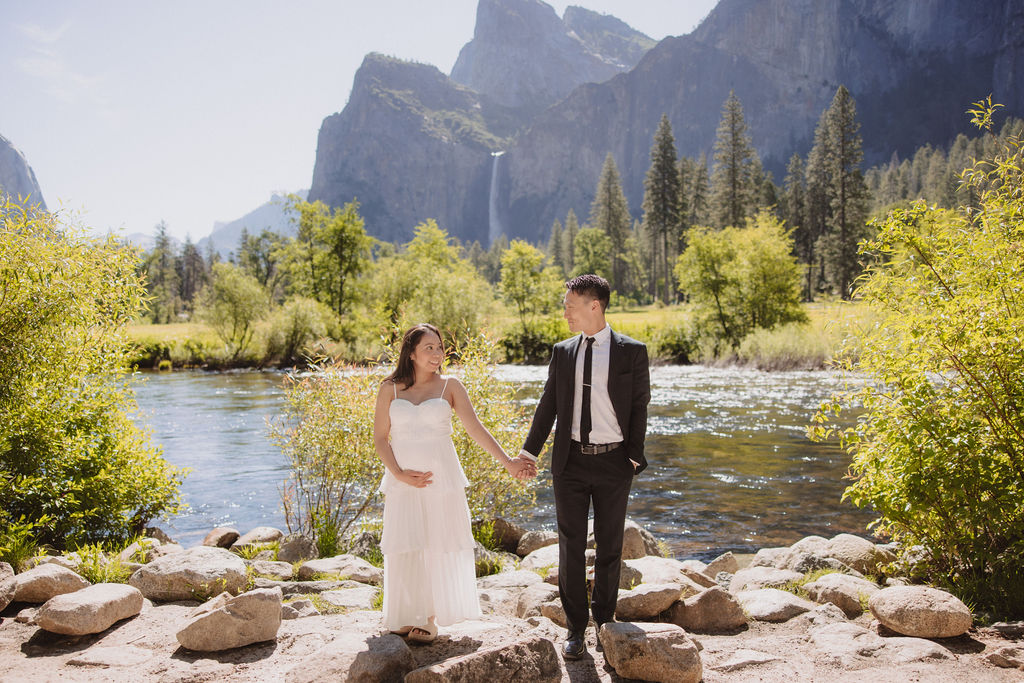 A couple in formal attire hold hands in a grassy field with a waterfall and towering cliffs in the background 