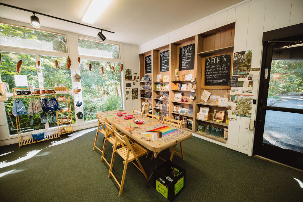 A well-lit activity room with a table set for art projects, surrounded by shelves and boards displaying art supplies, books, and information. Large windows and a door provide natural light.