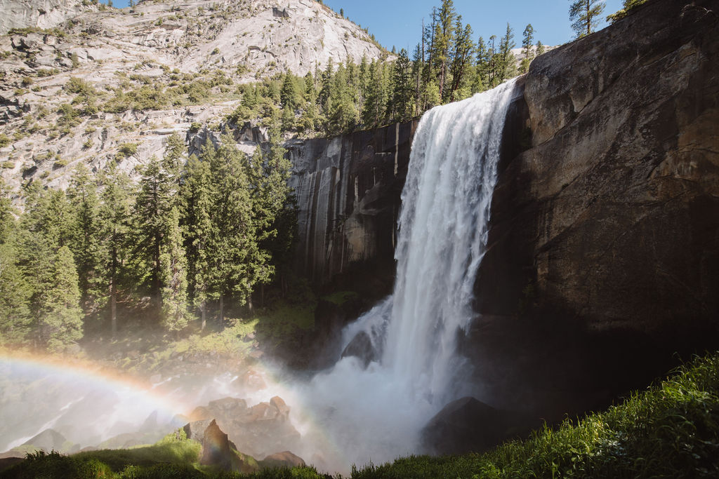 A tall waterfall cascades into a rocky pool surrounded by lush pine trees under a clear blue sky, with a rainbow visible in the mist at the base of the falls.