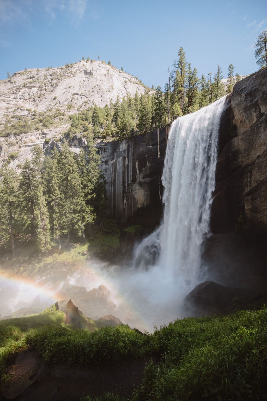 A tall waterfall cascades into a rocky pool surrounded by lush pine trees under a clear blue sky, with a rainbow visible in the mist at the base of the falls.