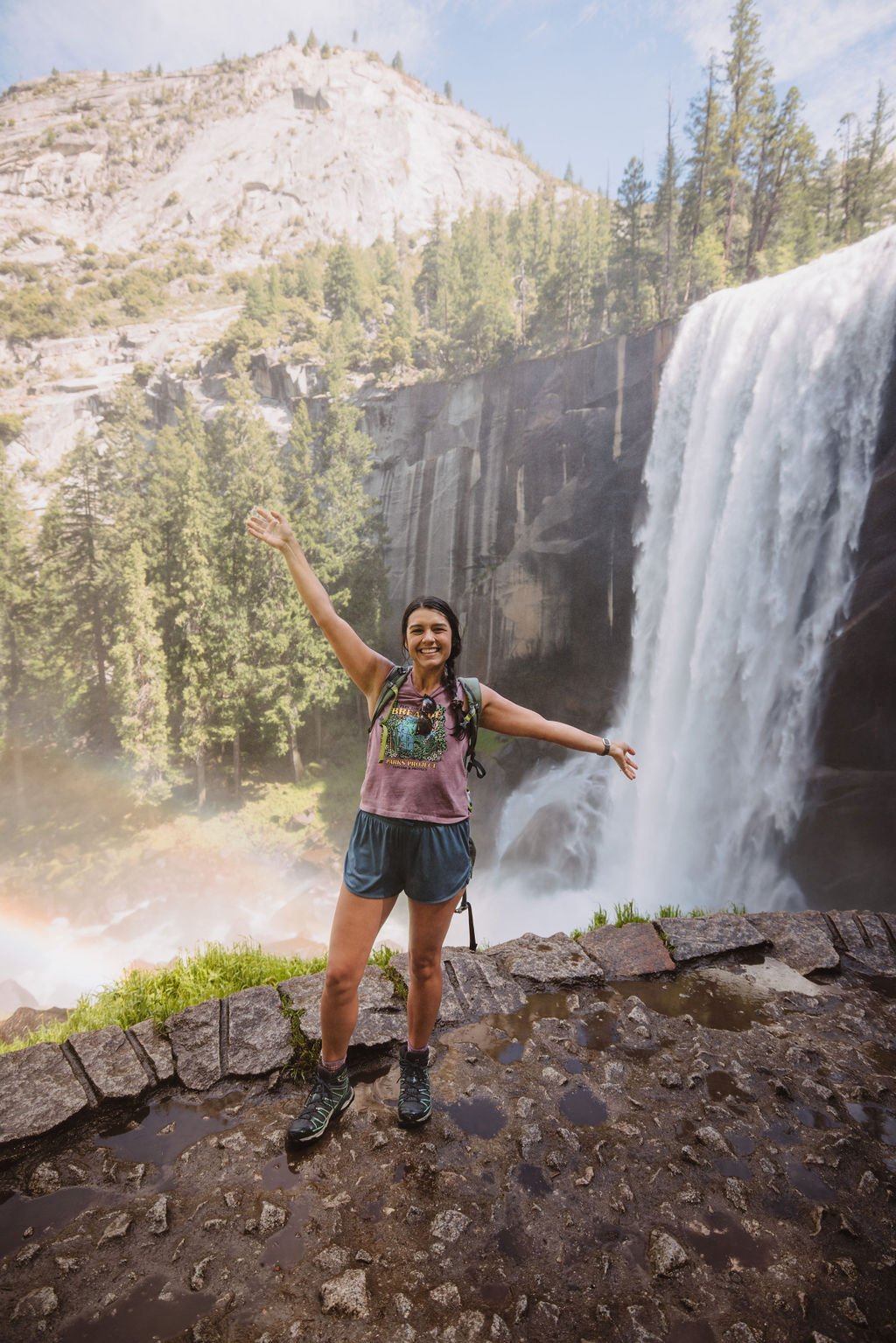 A person stands smiling with arms outstretched near a waterfall in a mountainous area. Trees and a rainbow are visible in the background.