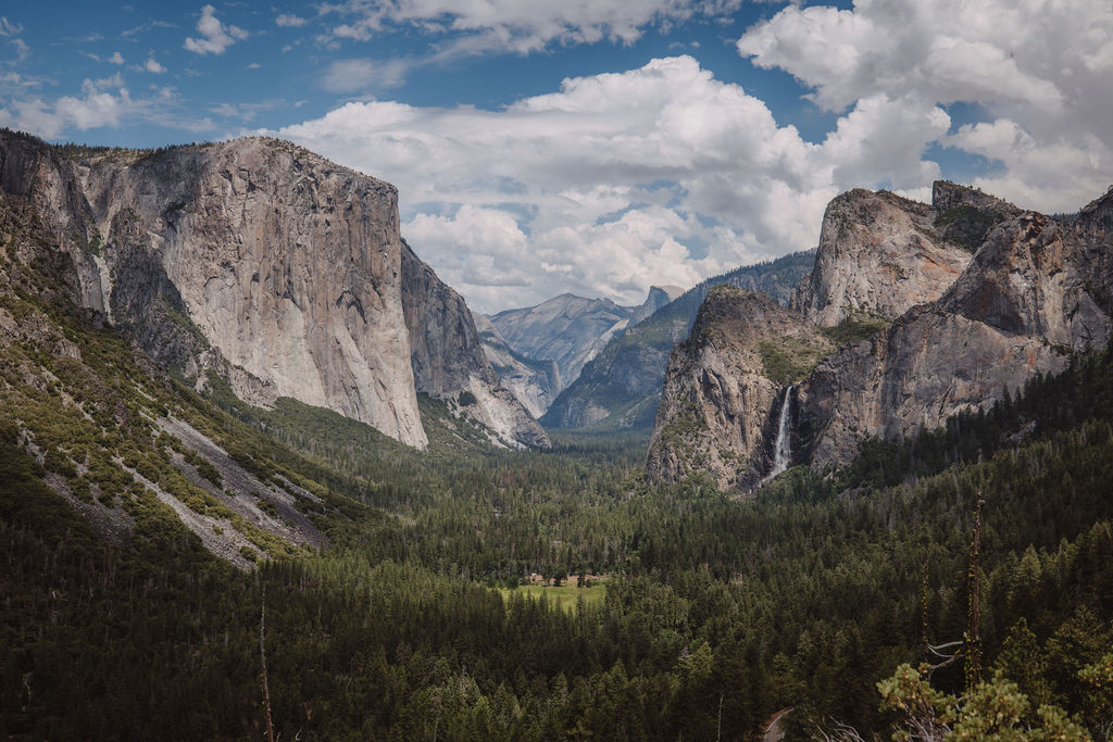 A scenic view of Yosemite Valley showcasing towering granite cliffs, green forested areas, and a distant range of mountains under a partly cloudy sky.