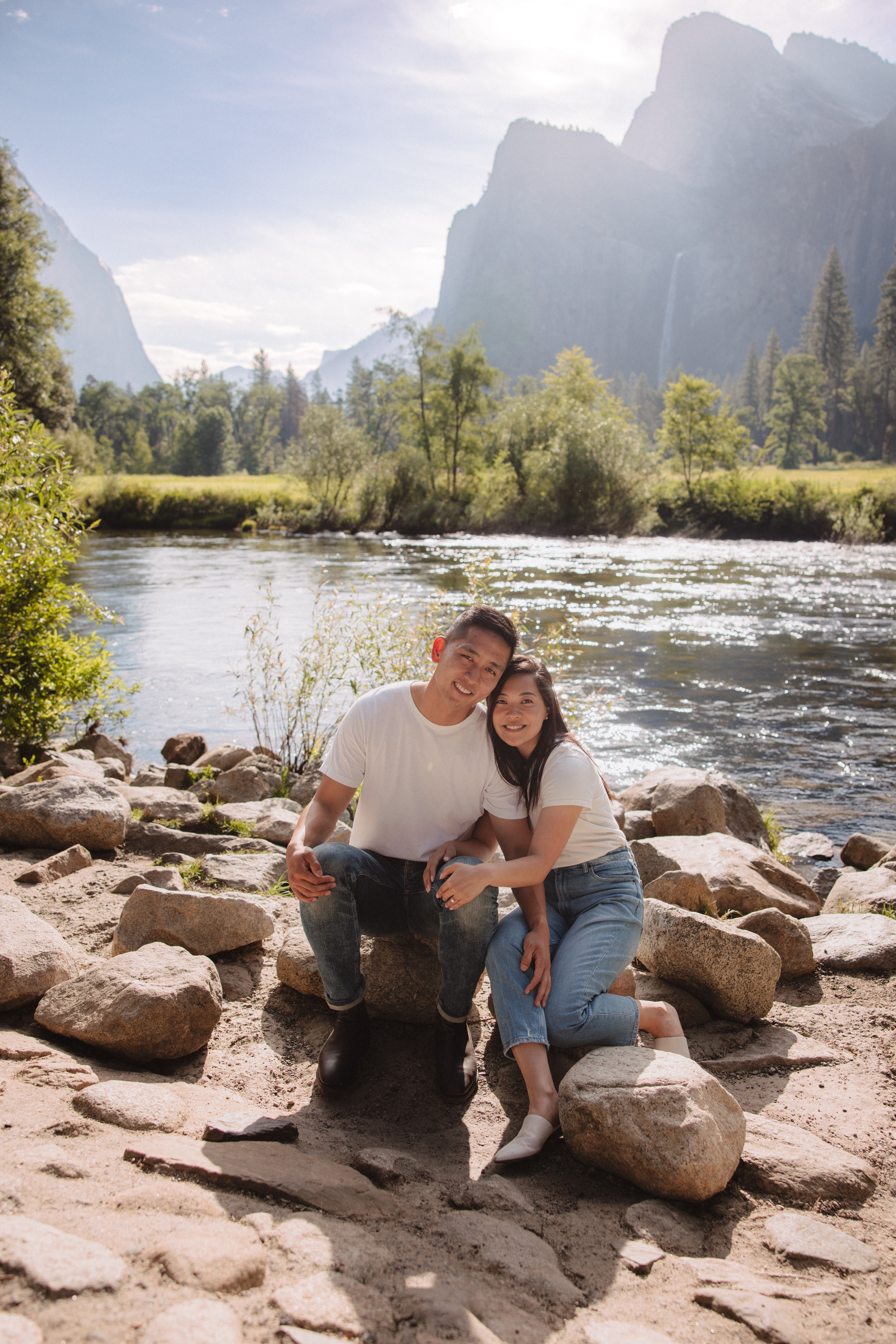 A couple sitting on rocks by a river in a scenic, mountainous landscape, both smiling and dressed in casual attire. The background features trees and distant mountains under a clear sky | Ultimate Yosemite Visitor Guide