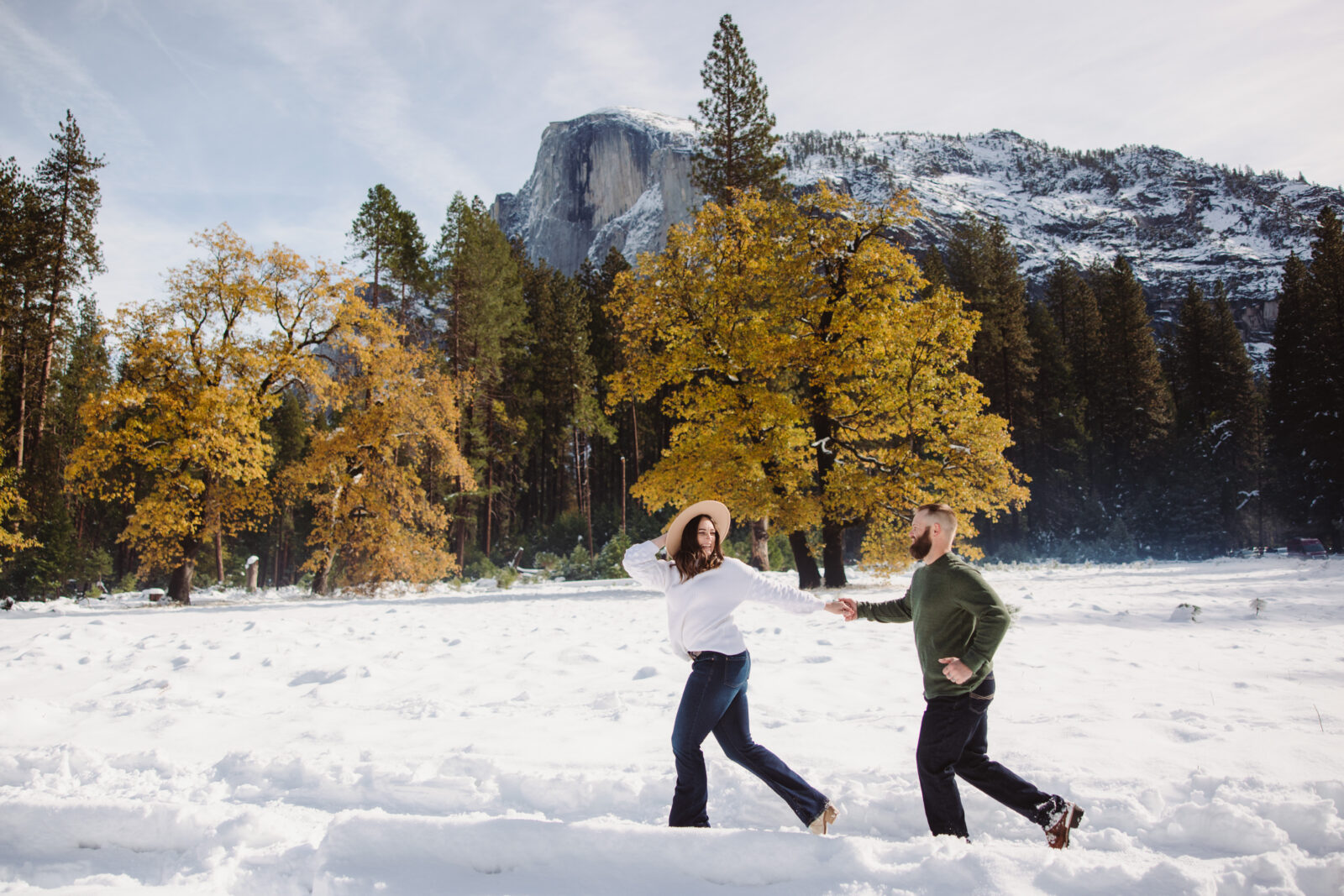 A couple walks hand in hand in a snowy field with tall trees and a mountain in the background. The woman wears a white sweater and hat, while the man wears a green sweater at half meadow dome | ultimate yosemite visitor guide