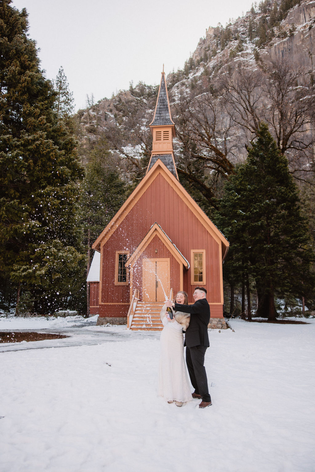 A couple in wedding attire stands in front of a small chapel with snow-covered ground and trees around. The man is popping a bottle of champagne. Ultimate Yosemite Visitor Guide 