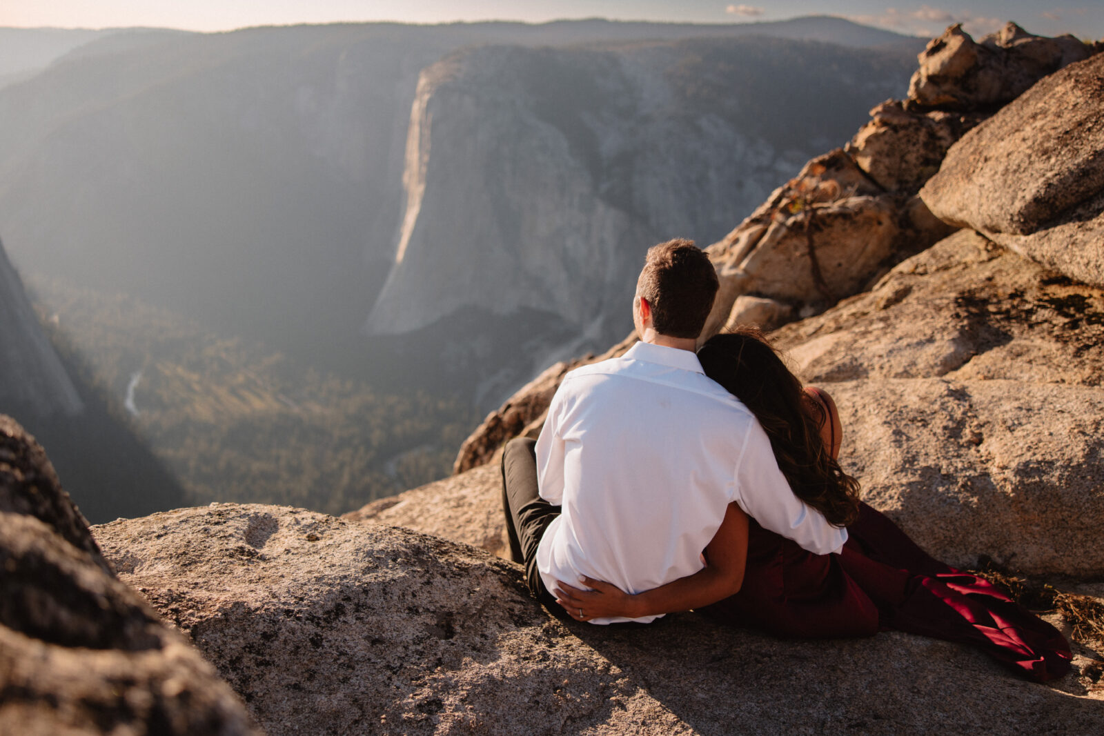 A couple sits on a rocky ledge, with the man's arm around the woman, overlooking a valley and distant mountains at sunset at taft point | Ultimate Yosemite Visitor Guide 