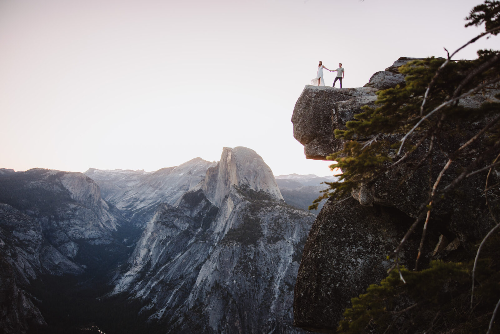 Two people stand holding hands on the edge of a cliff with a mountainous landscape in the background during sunset at Glacier point in Yosemite National park | Ultimate yosemite visitor guide