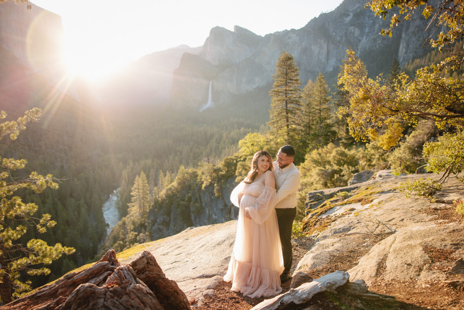 A couple embraces on a rocky ledge, surrounded by trees and mountains, with the sun setting in the background at Tunnel View | Ultimate Yosemite Visitor Guide