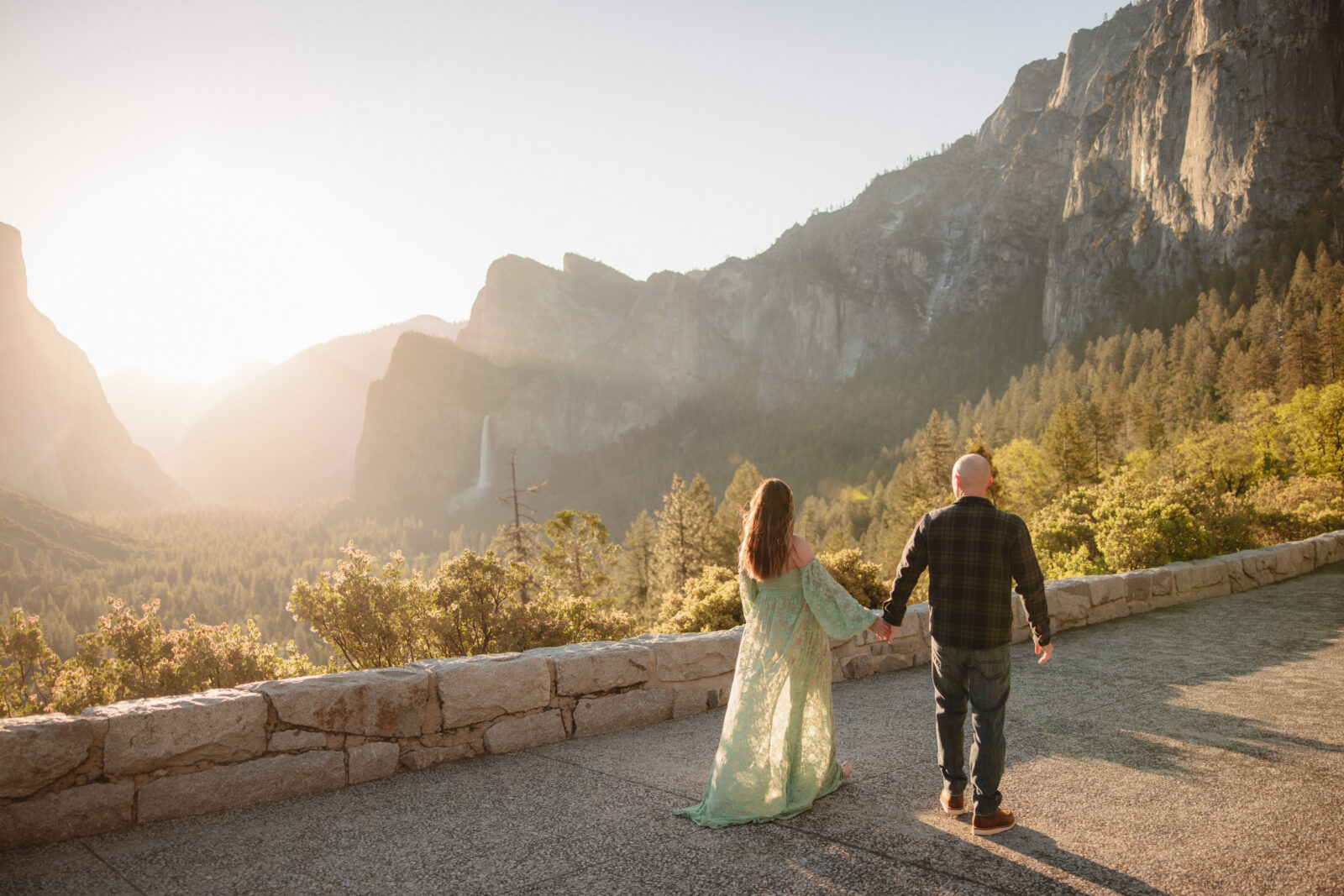 A couple embraces on a rocky ledge, surrounded by trees and mountains, with the sun setting in the background at Tunnel View | Ultimate Yosemite Visitor Guide