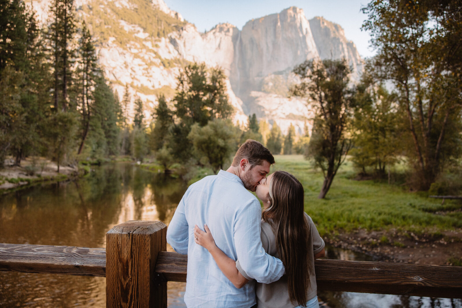 A couple stands on a wooden bridge, kissing and embracing, with a serene river and a towering mountain in the background. The scene is set in a lush, green valley surrounded by trees at Swinging bridge meadow | Ultimate Yosemite Visitor Guide 