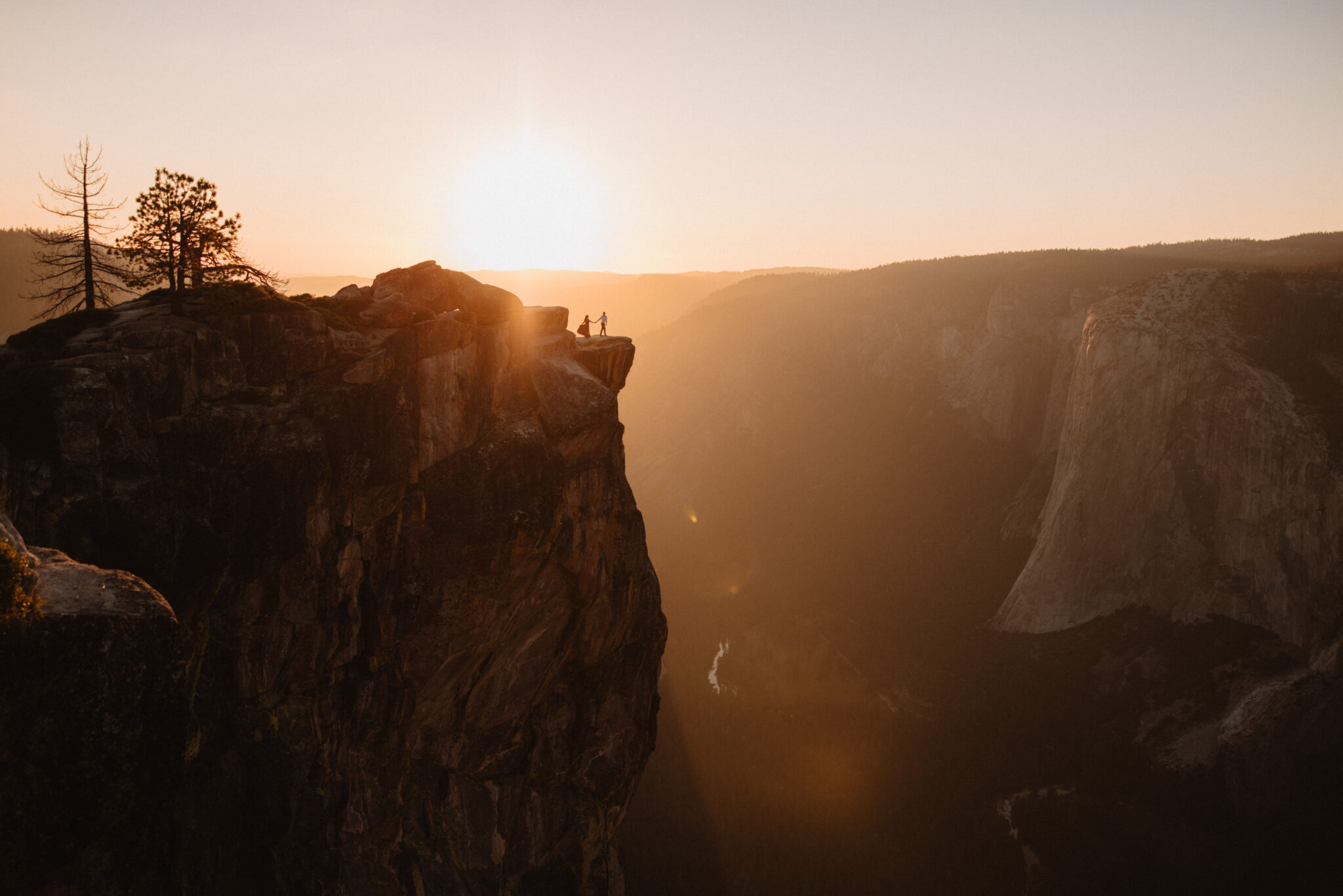 Two people stand on a cliff edge at sunrise in a mountainous area with a valley below and another mountain in the distance at Taft Point at Yosemite national park | Ultimate Yosemite visitor guide