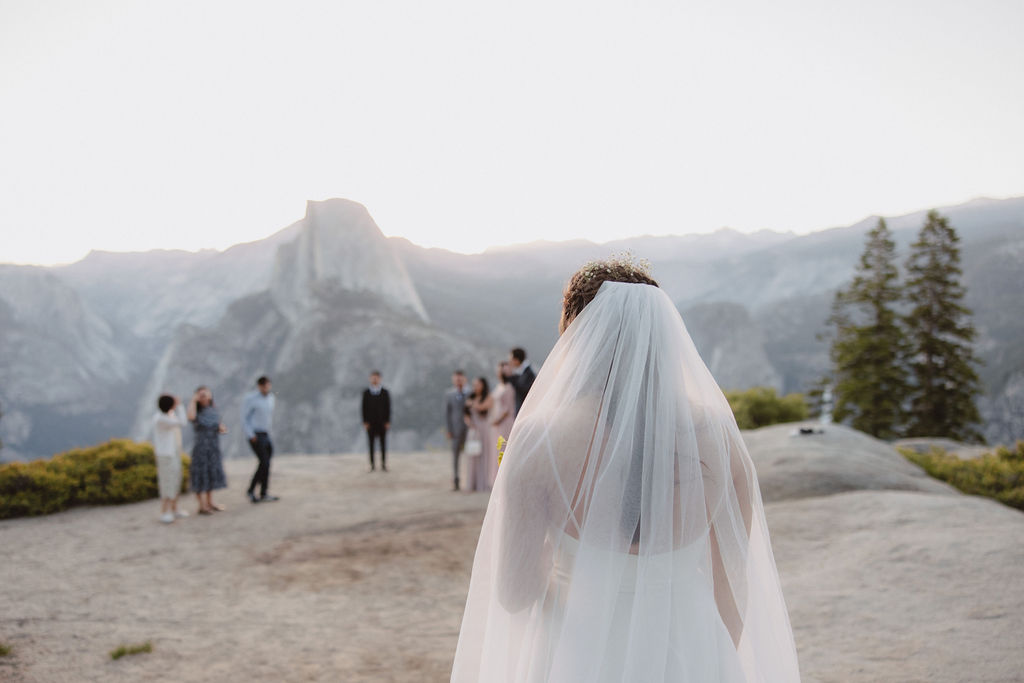 A person in a wedding veil faces a group of people standing on a scenic overlook with mountains in the background during a sunrise ceremony in Yosemite