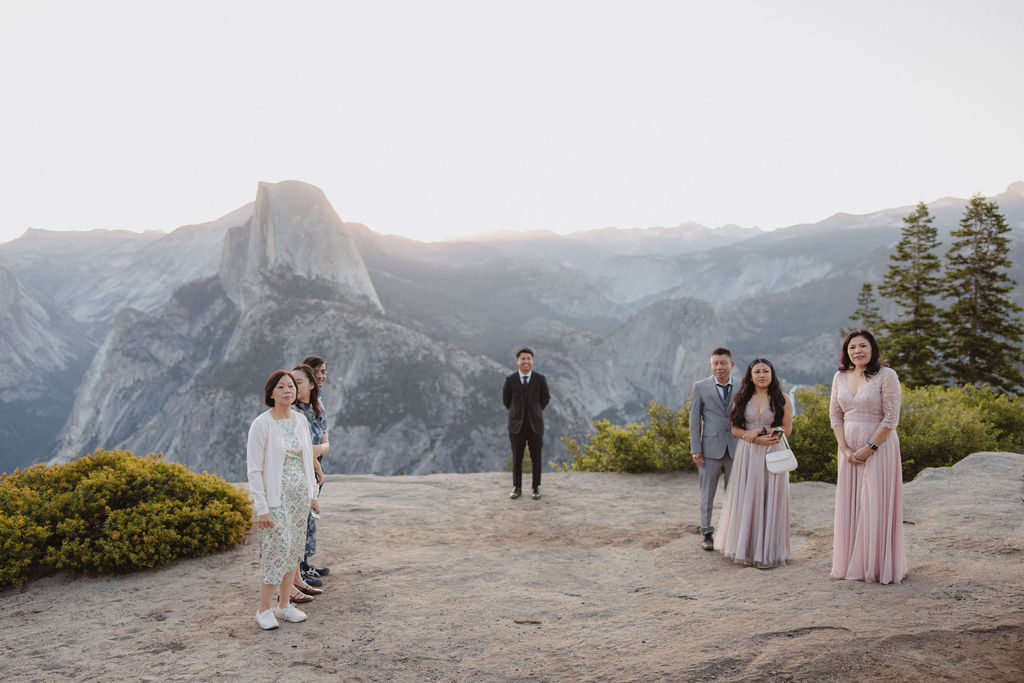 A person in a wedding veil faces a group of people standing on a scenic overlook with mountains in the background during a sunrise ceremony in Yosemite