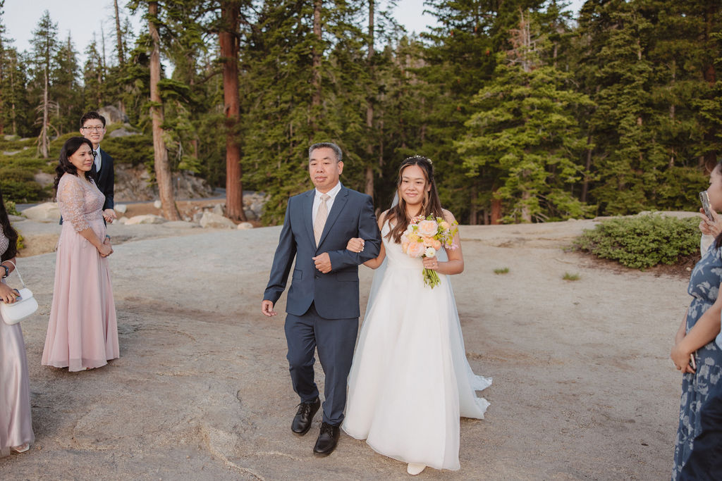 A person in a wedding veil faces a group of people standing on a scenic overlook with mountains in the background during a sunrise ceremony in Yosemite