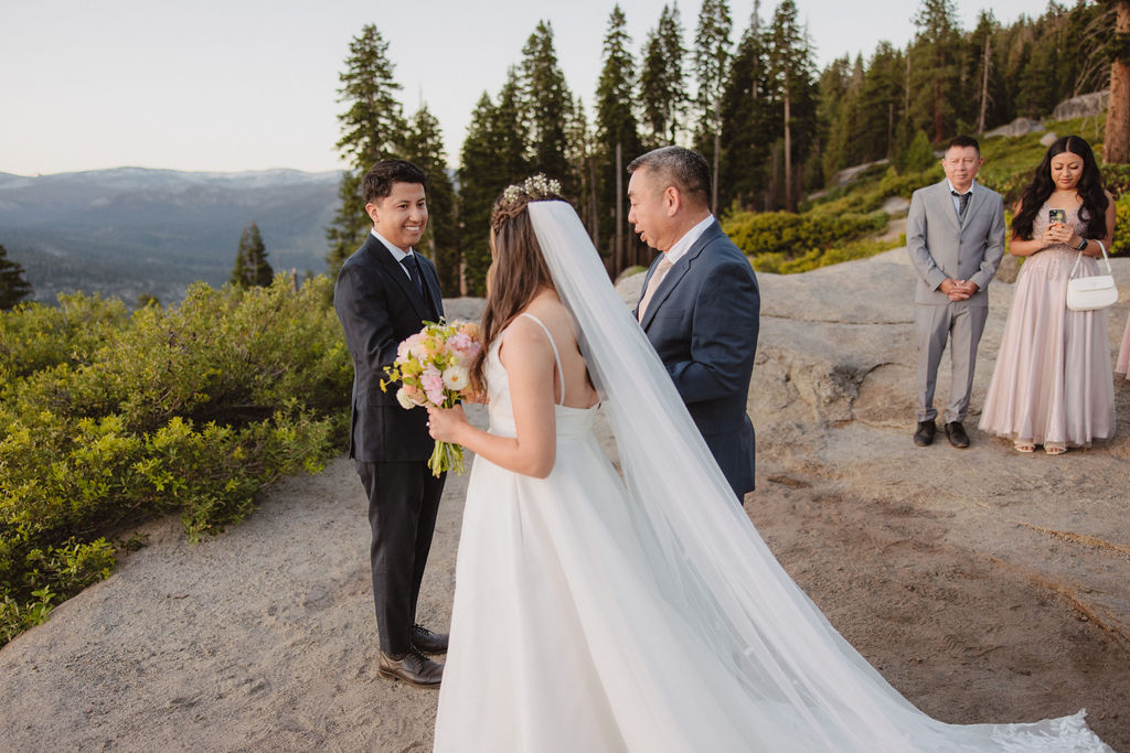 A person in a wedding veil faces a group of people standing on a scenic overlook with mountains in the background during a sunrise ceremony in Yosemite