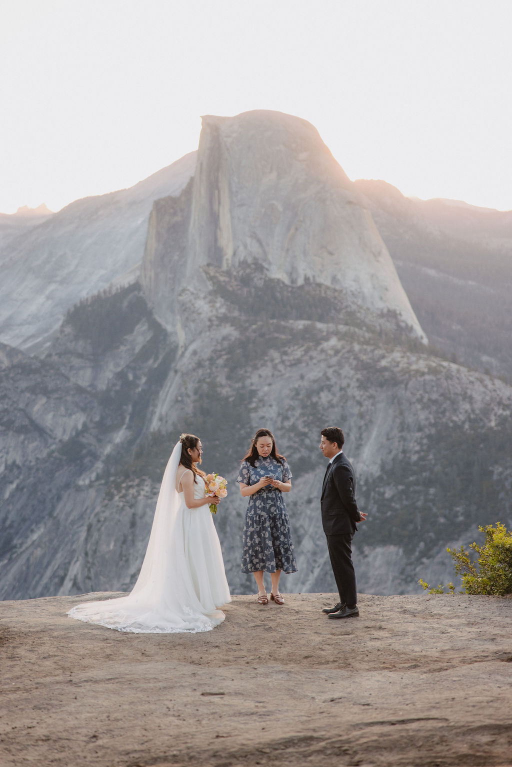 Bride and groom in wedding attire stand on a rocky ledge with an officiant in front of them. A large mountain is in the background.