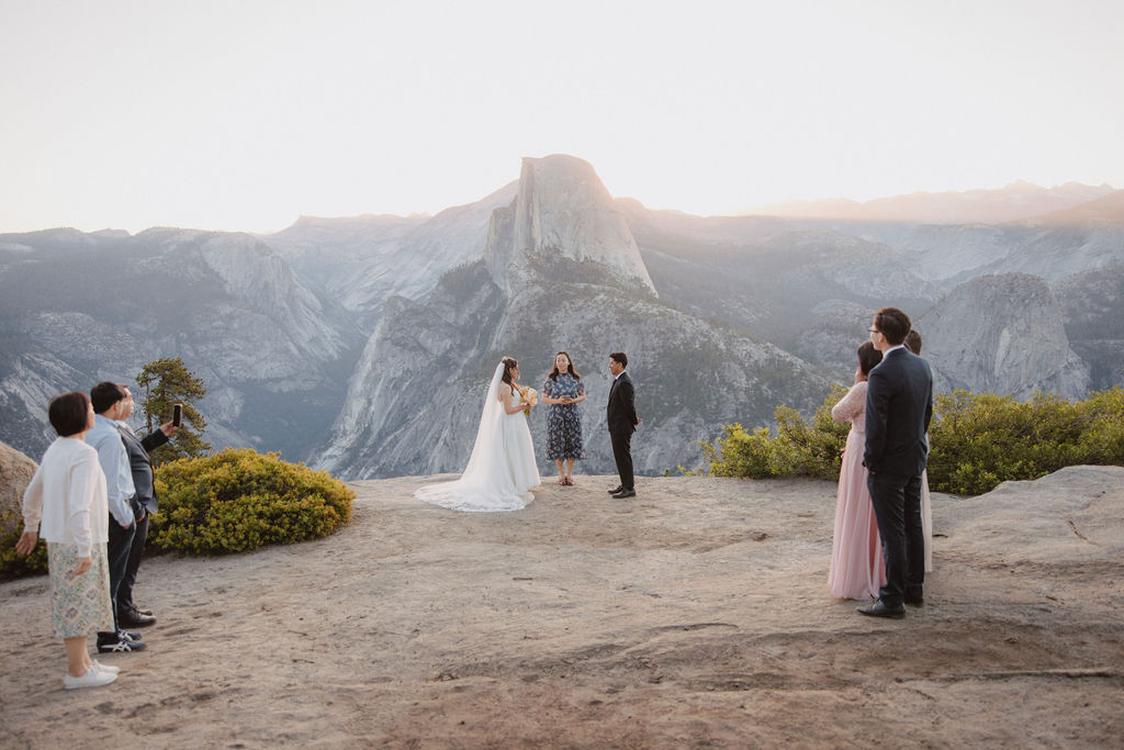 A bride and groom stand facing each other with an officiant between them during an outdoor wedding ceremony with a large mountain in the background in Yosemite