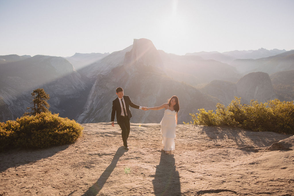 A man lifts a woman in his arms as they pose on a sunny mountain overlook with rugged peaks and trees in the background at their elopement in yosemite