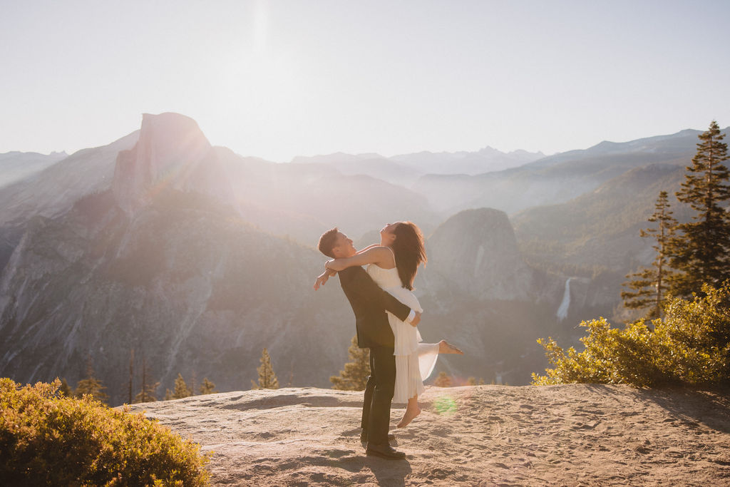 A man lifts a woman in his arms as they pose on a sunny mountain overlook with rugged peaks and trees in the background at their elopement in yosemite