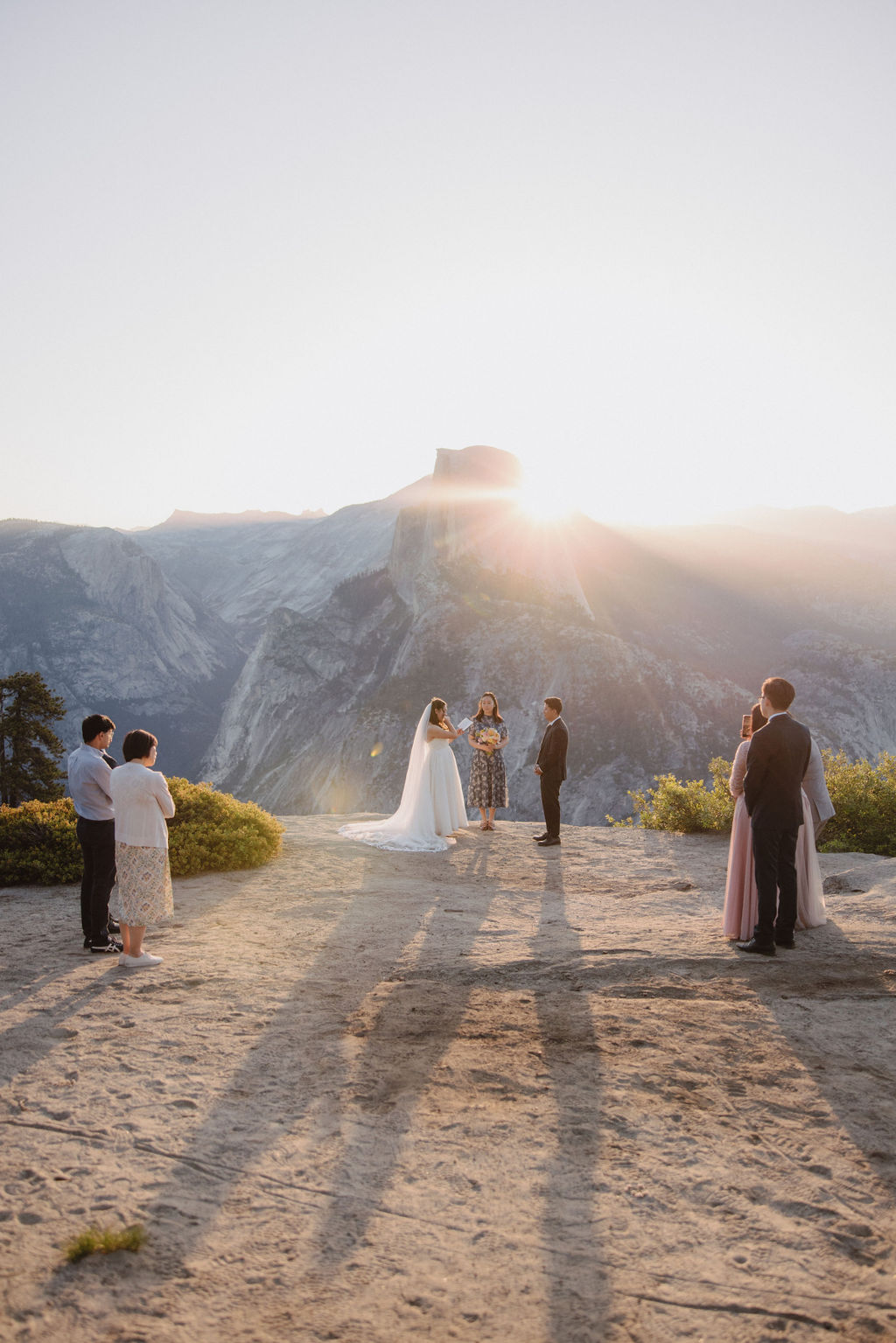 A couple stands with an officiant and wedding party on a rocky outcrop with a sunlit mountain in the background, as three pairs of guests look on in Yosemite 